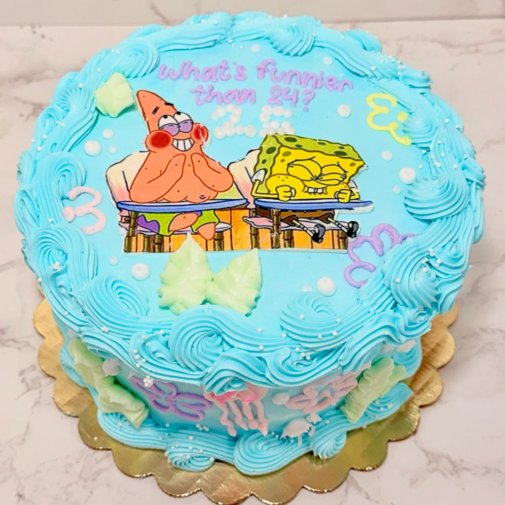 A round birthday cake decorated with light blue frosting, featuring SpongeBob and Patrick illustrations, colorful text, and piped swirls on top and sides, set on a gold scalloped cake board against a light marble background.
